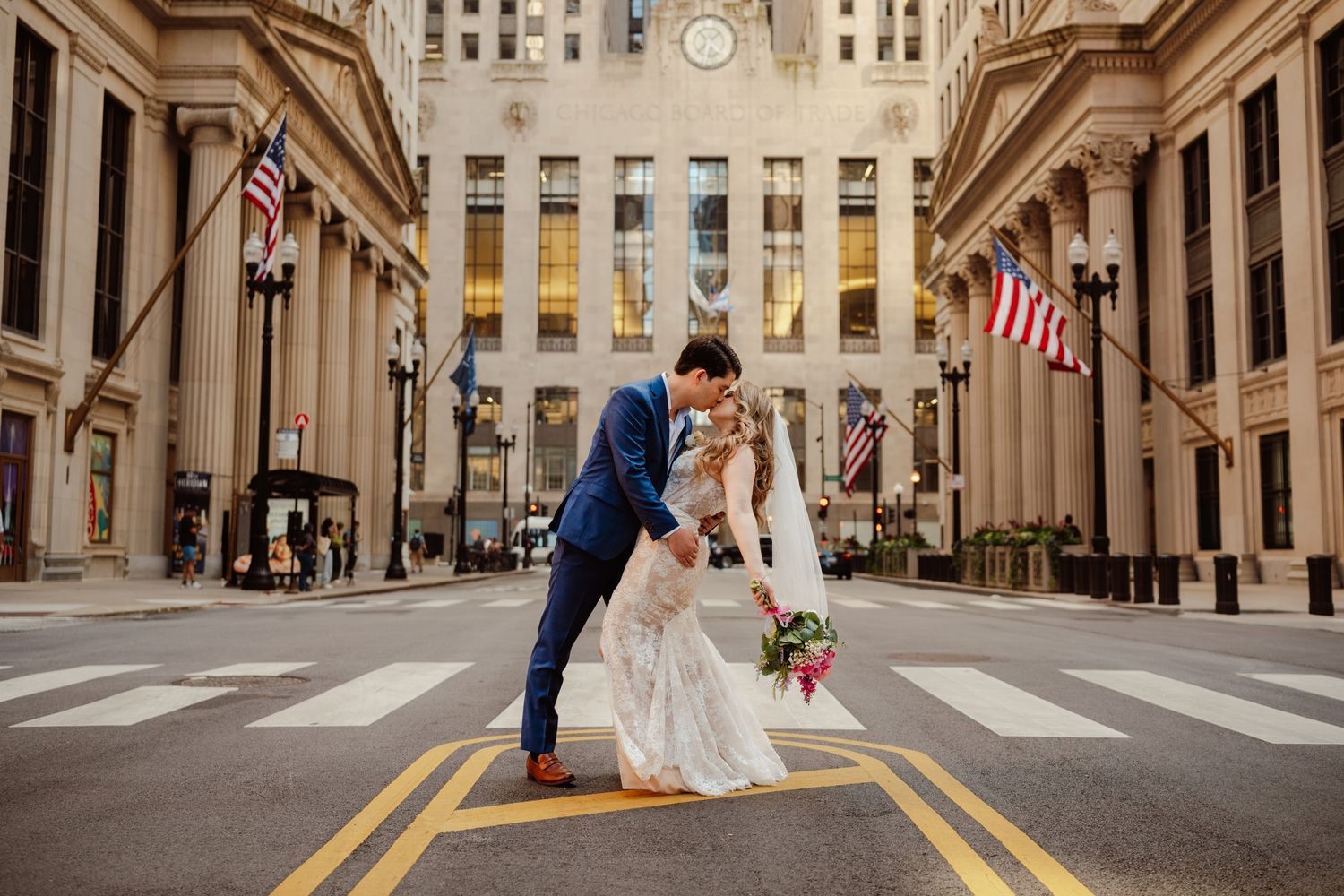 Bride and groom share a romantic dip kiss on a city street crosswalk with grand classical architecture and American flags.