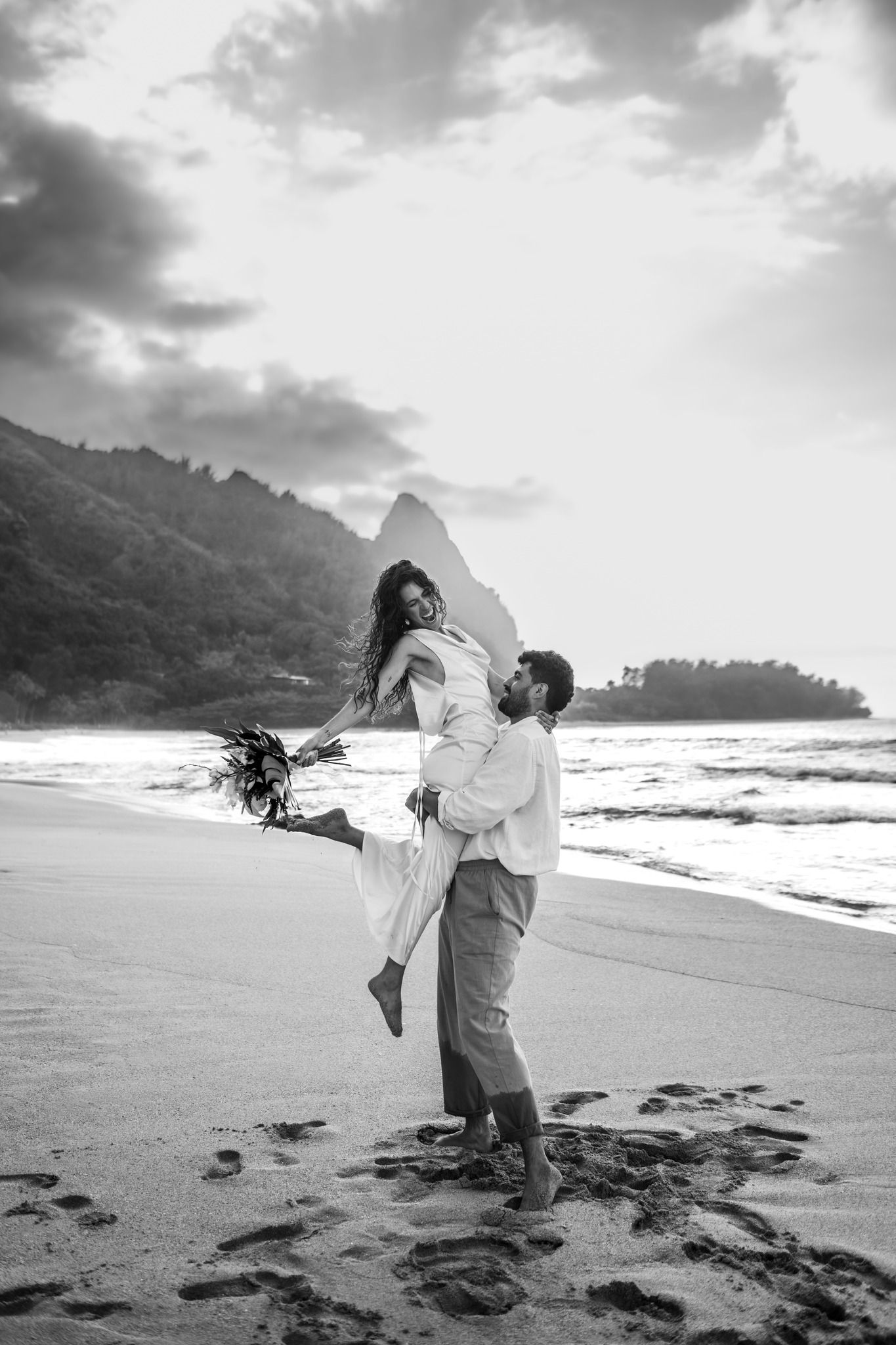 Black and white photo of groom lifting bride on a beach at sunset in Hawaii.