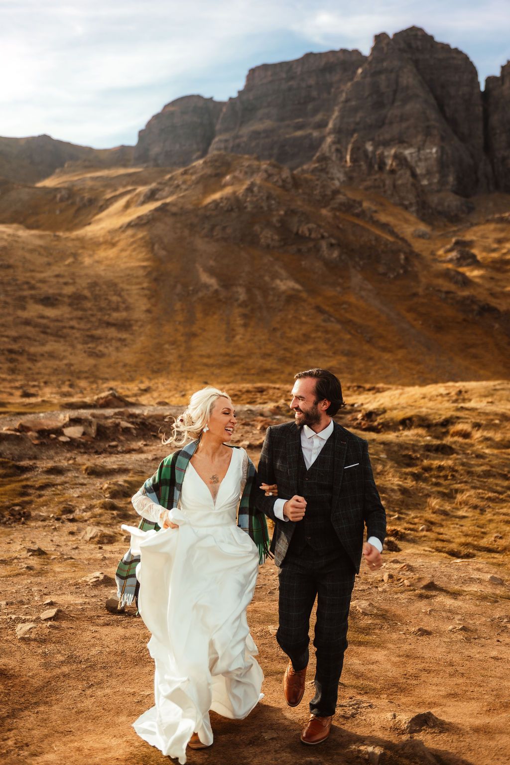 Bride and groom walk hand-in-hand through rugged Scottish highlands landscape in wedding attire.