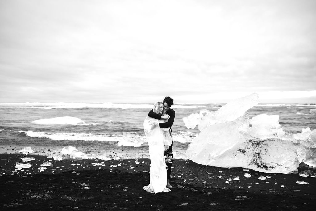 Couple embraces on black sand beach with icebergs in Iceland elopement wedding photo.