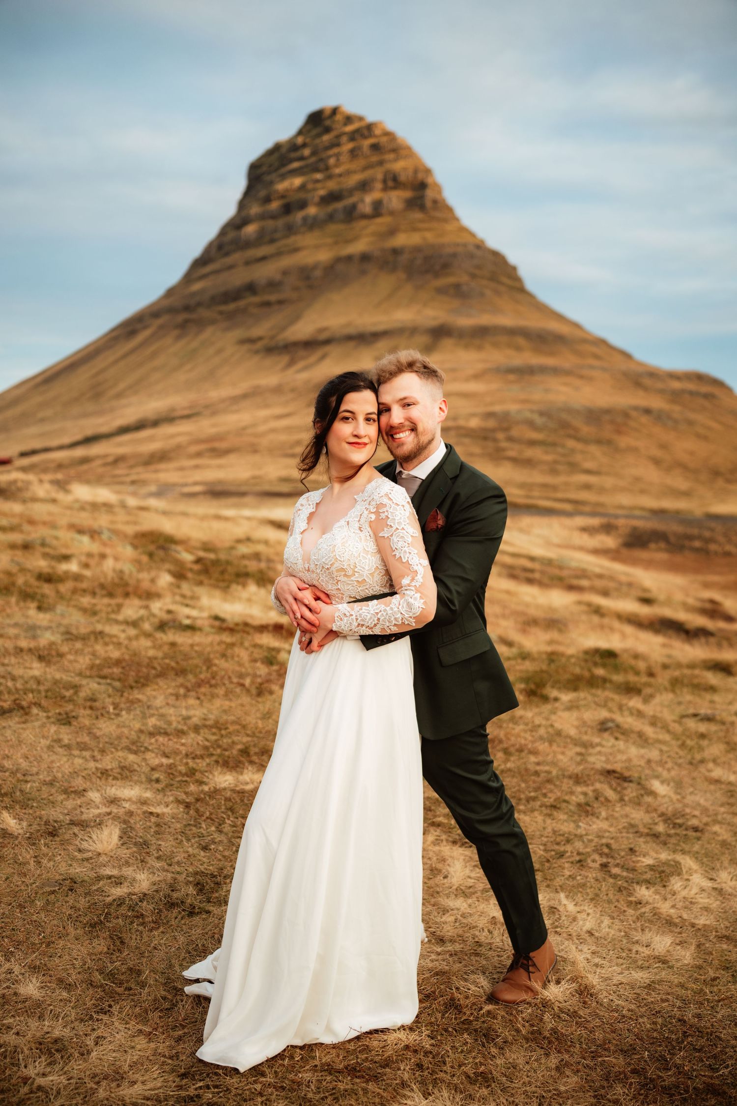 A bride and groom embrace before Kirkjufell mountain in Iceland, she in white lace gown, he in dark suit.