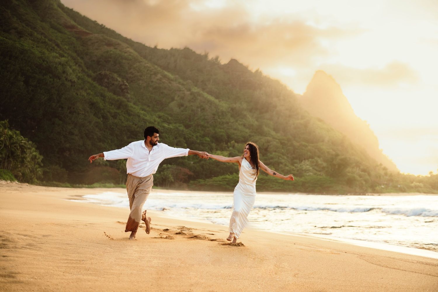 Couple holding hands running on a golden sandy beach at sunset with lush green mountains in the background.