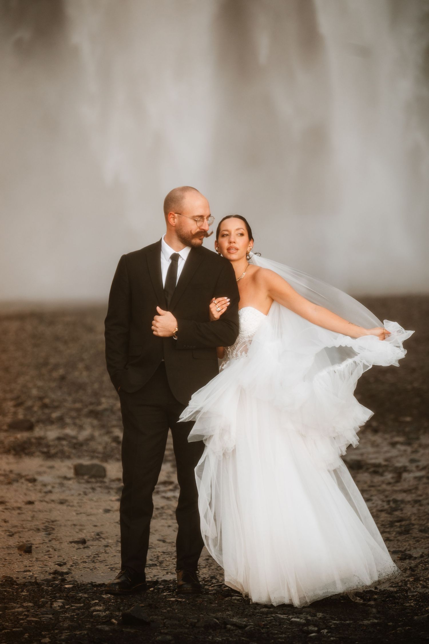 Bride in flowing white gown and veil poses with groom in black suit before dramatic Icelandic waterfall backdrop.