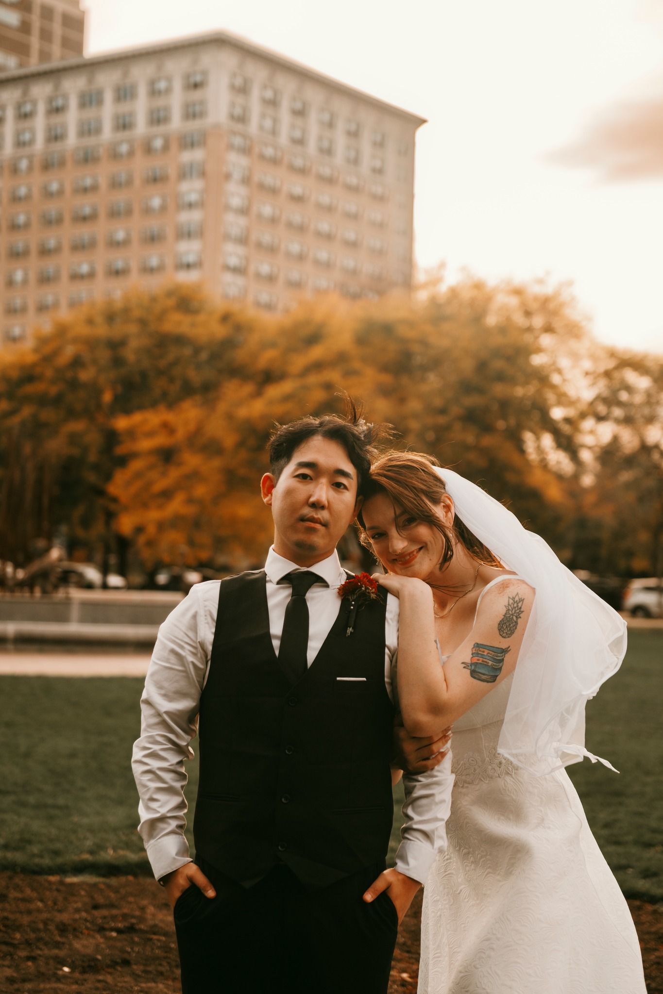 Asian groom in black vest and bride in white dress posing together in urban park with autumn foliage backdrop.