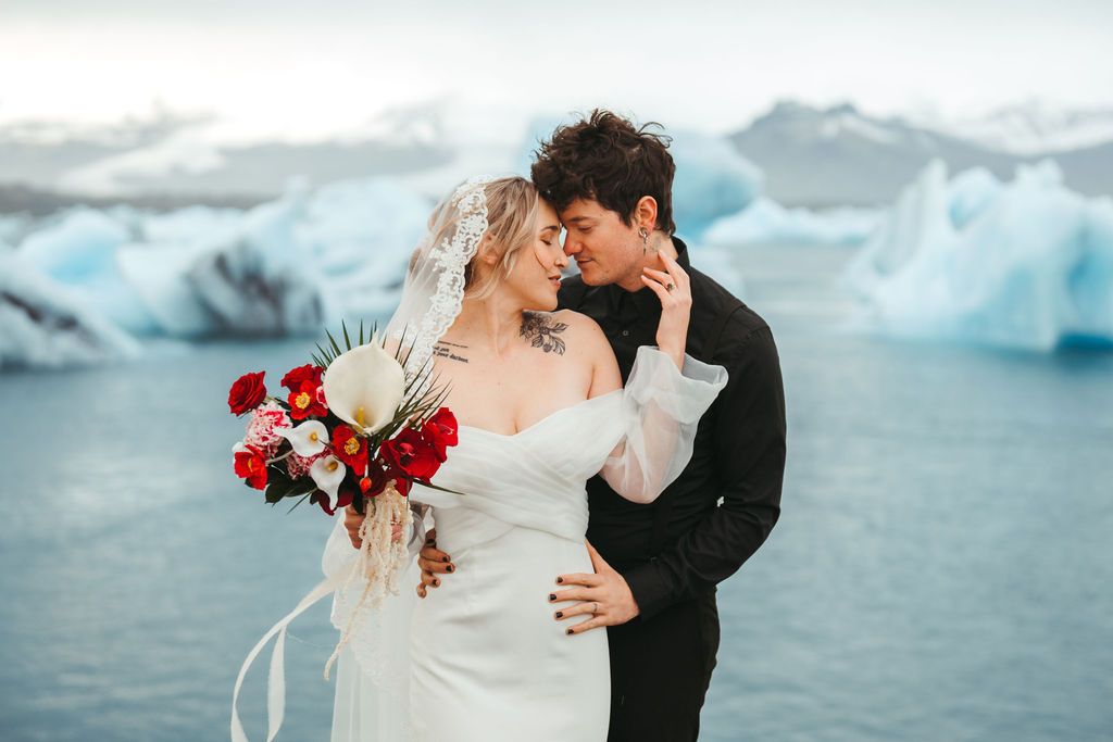 Couple sharing a romantic moment at a glacier lagoon wedding in Iceland, bride holding a red bouquet.