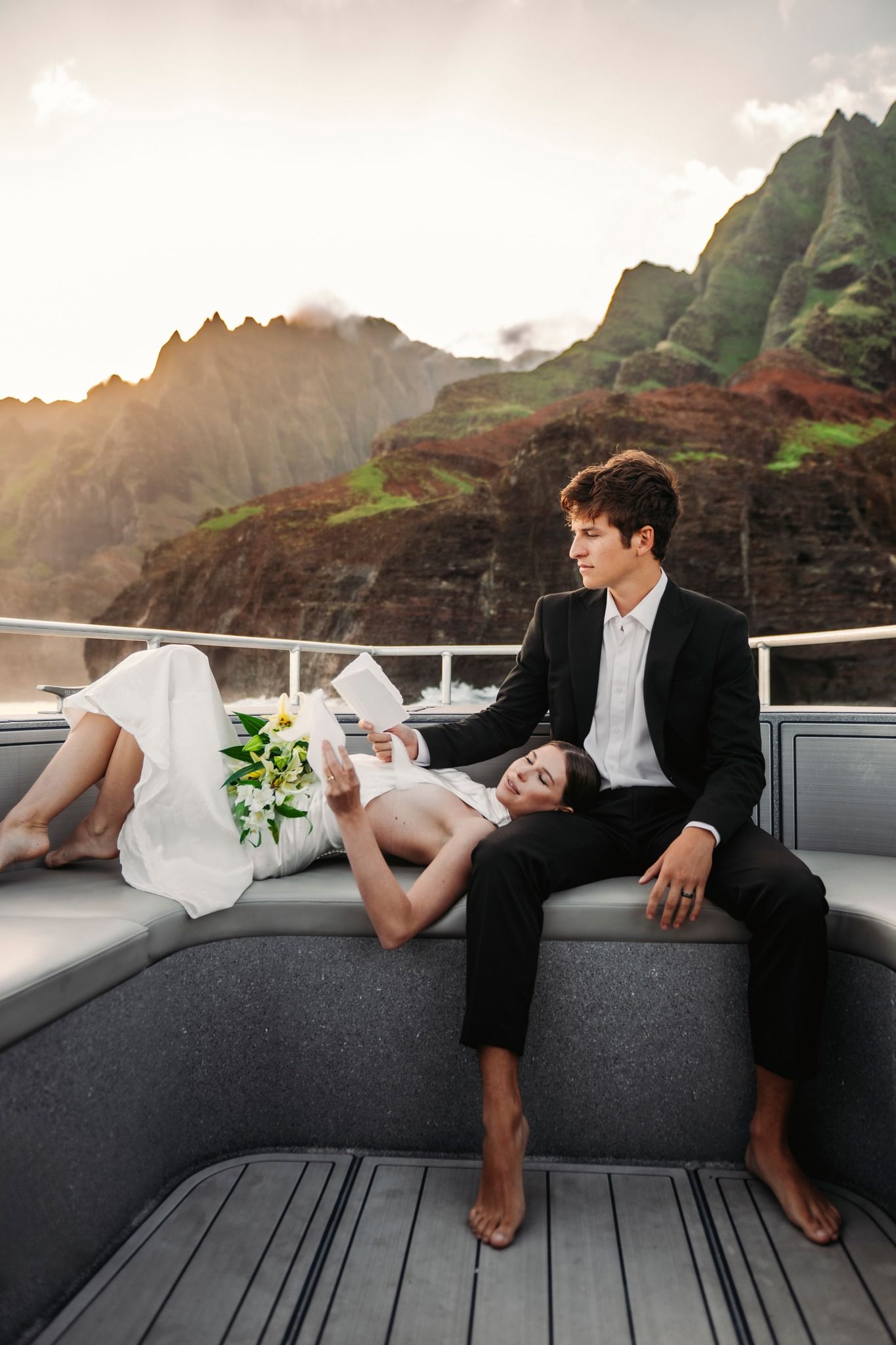 Bride and groom in wedding attire relaxing on a yacht with dramatic Na Pali Coast mountains at sunset in Hawaii.