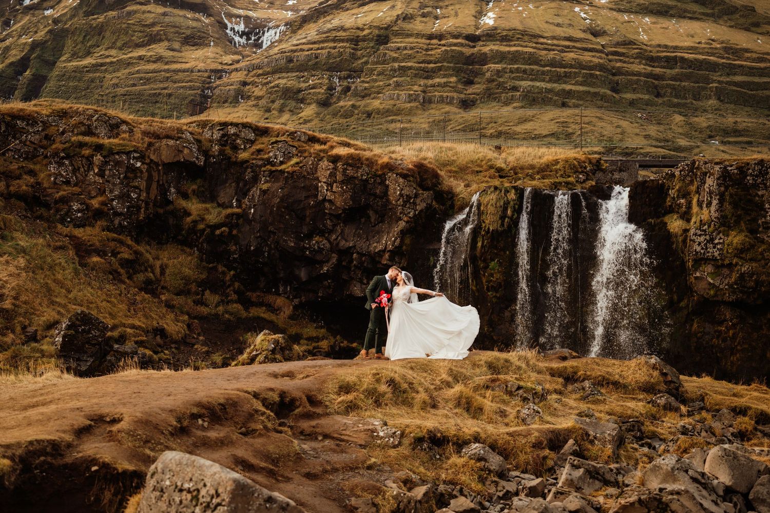 Bride and groom embrace near a stunning waterfall in Iceland's dramatic green mountain landscape.