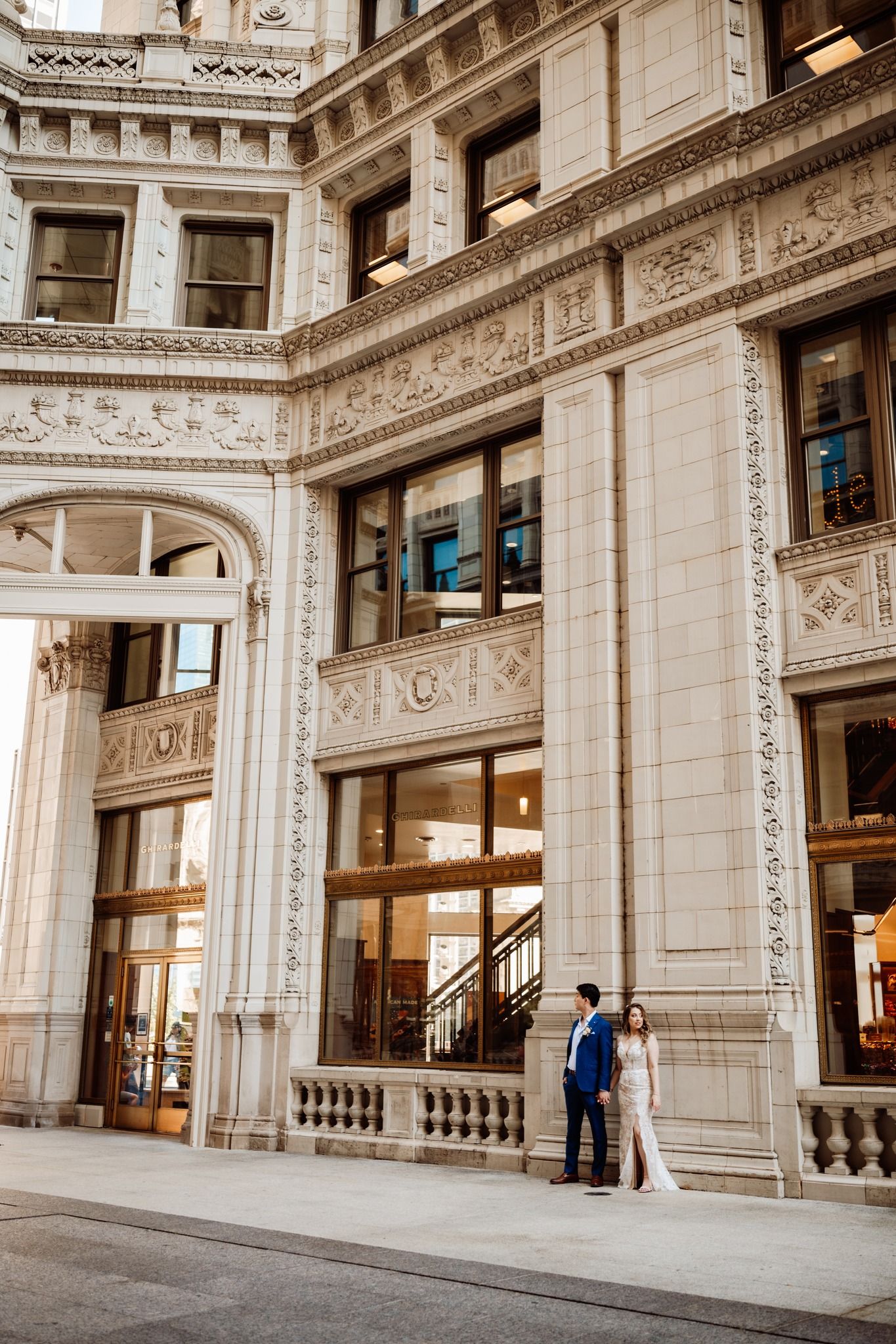 Bride and groom pose outside an ornate beige stone building with arched windows and decorative carvings.