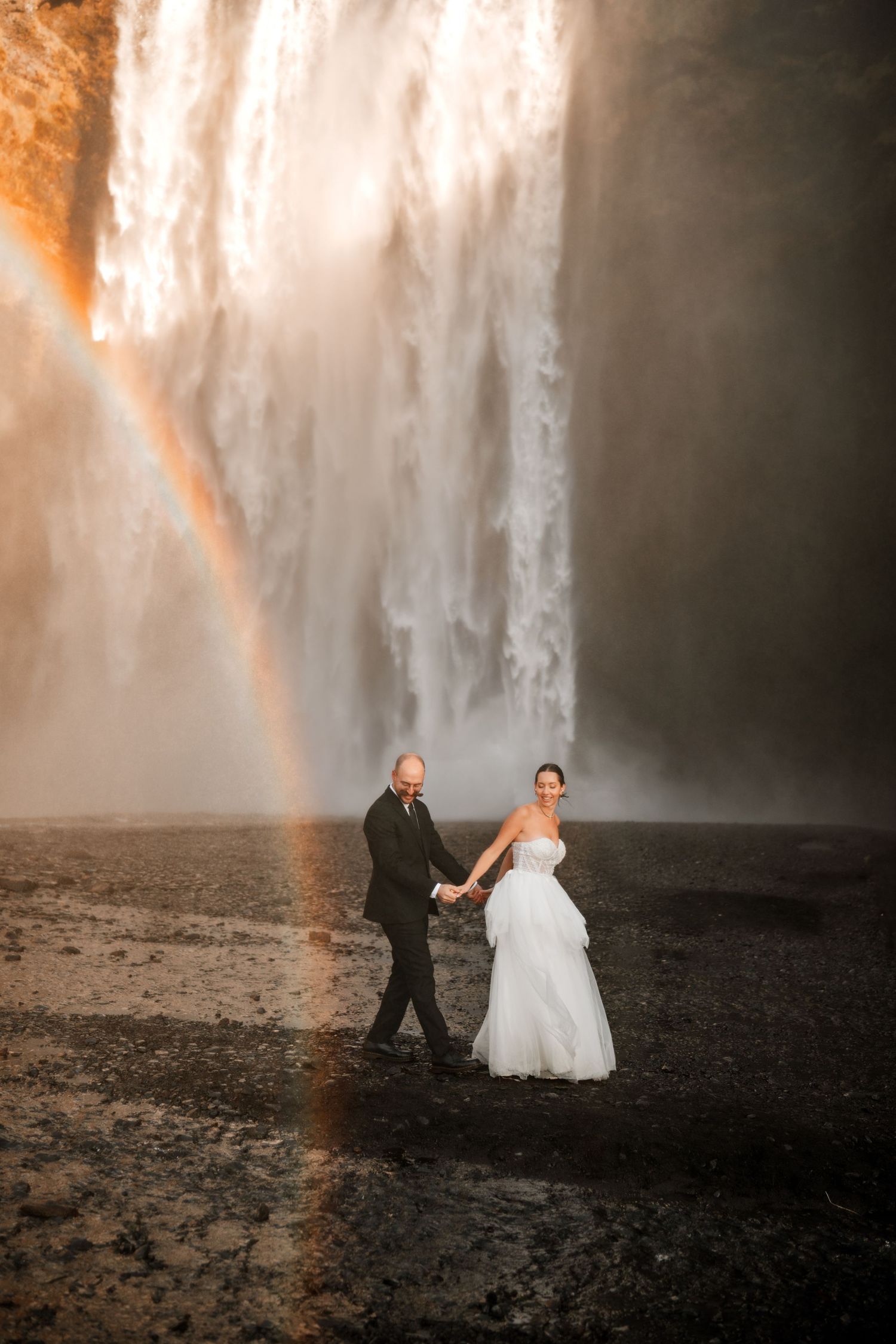 Bride and groom hold hands before a stunning waterfall with a rainbow arc in Iceland.