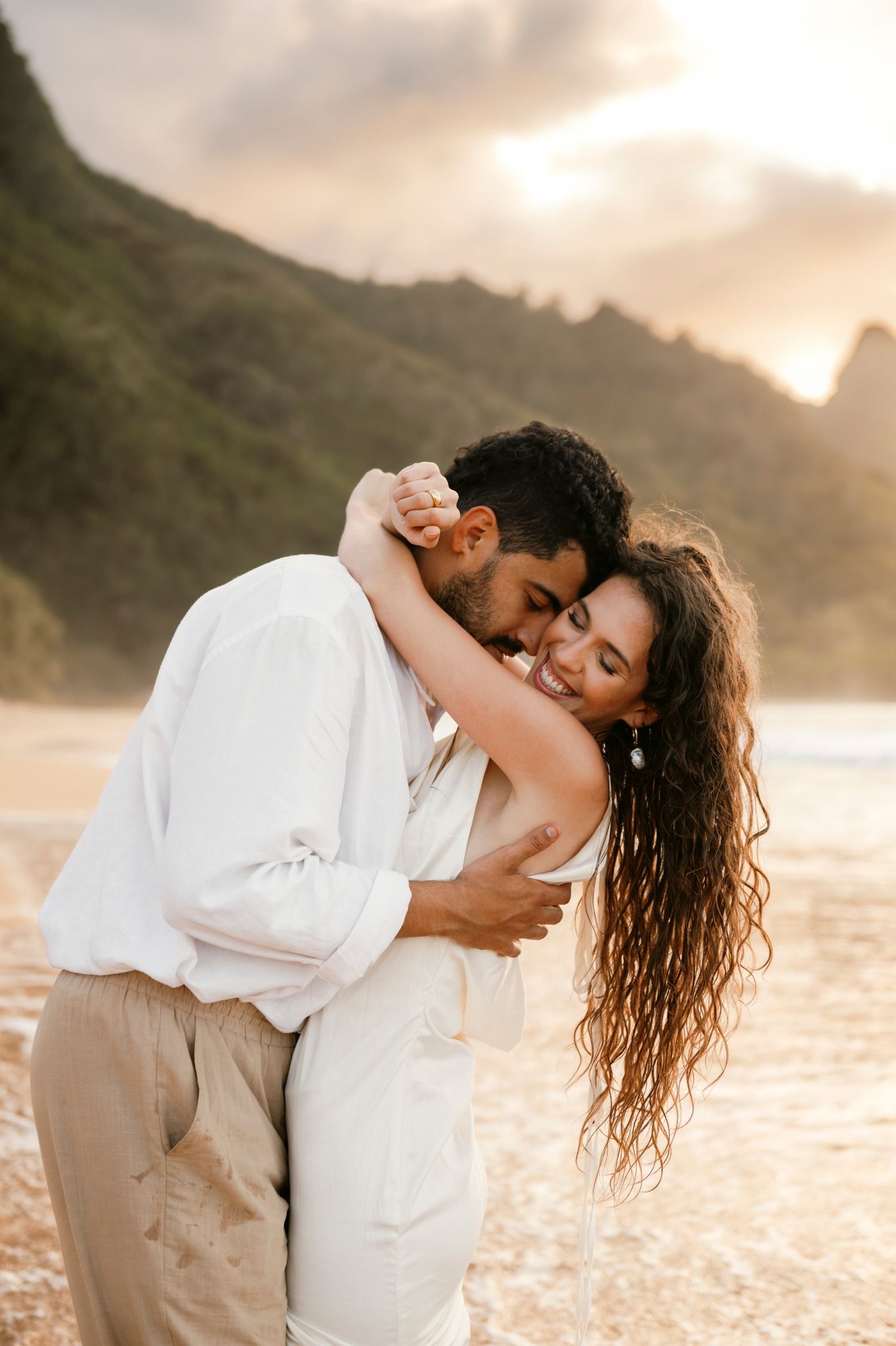 Couple embracing on a golden sunset beach with lush green mountains in the background.