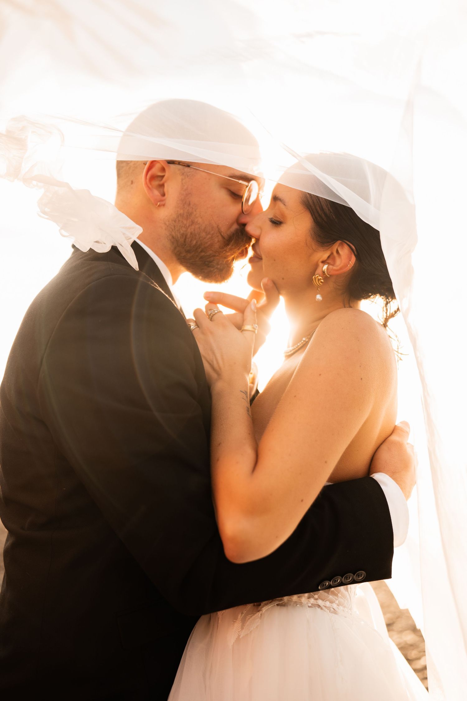 Bride and groom sharing a romantic kiss under a white veil with warm golden backlight at their wedding.