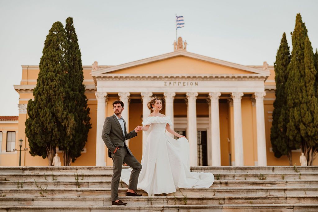 Bride and groom holding hands on steps of Zappeion in Athens, Greece, with cypress trees and Greek flag.