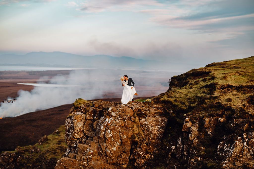 Bride and groom embrace on rocky cliff edge with dramatic misty landscape and cloudy sky in Iceland.