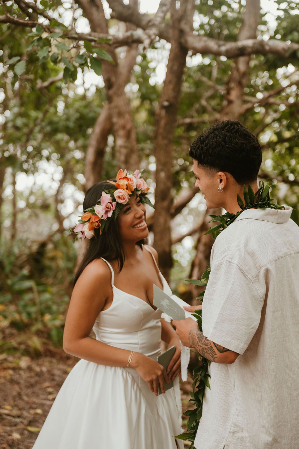 Bride in white dress and floral crown exchanging vows with groom in tropical forest setting.