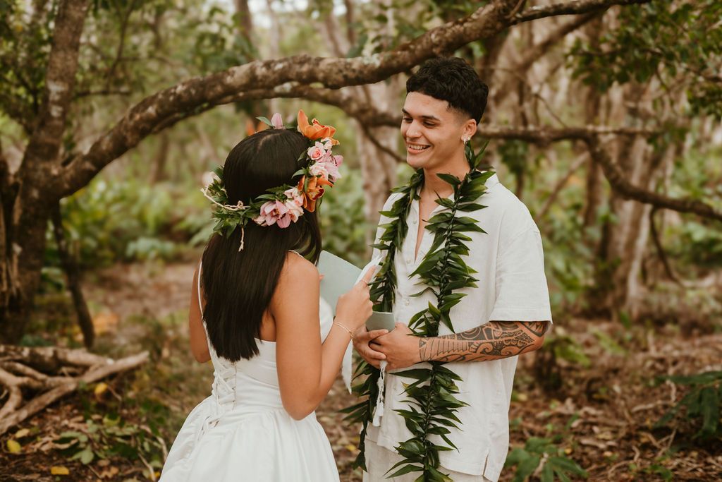 Groom with green lei smiling at bride wearing floral crown in tropical forest setting.