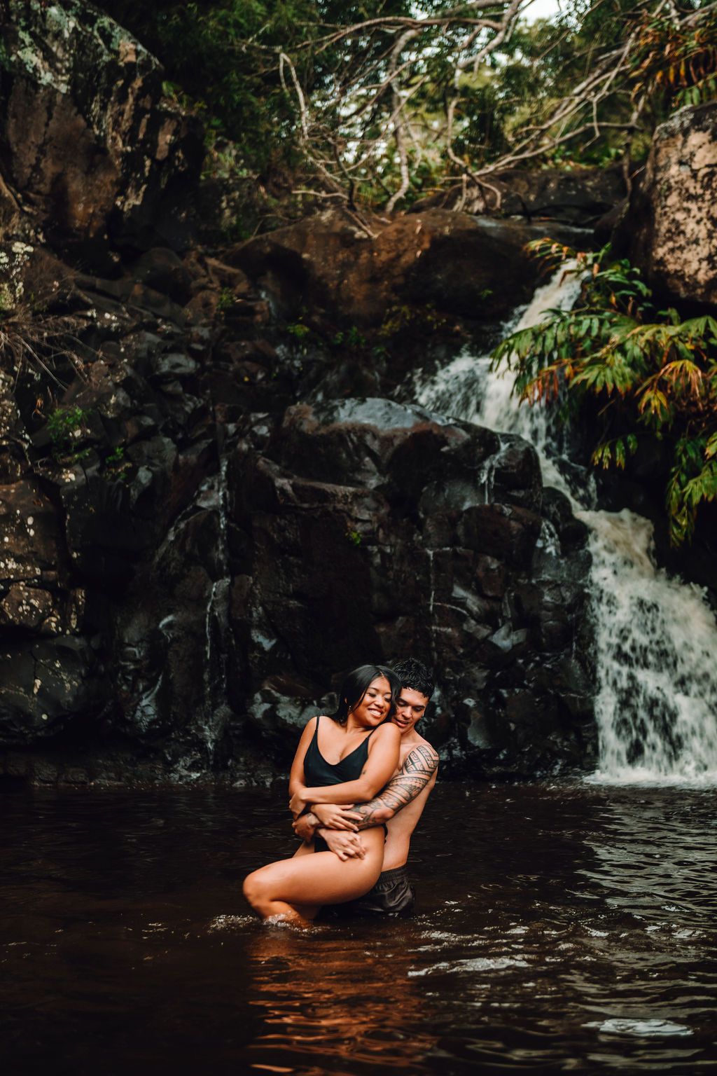 Couple embracing romantically in a tropical waterfall pool surrounded by lush greenery and dark rocks.