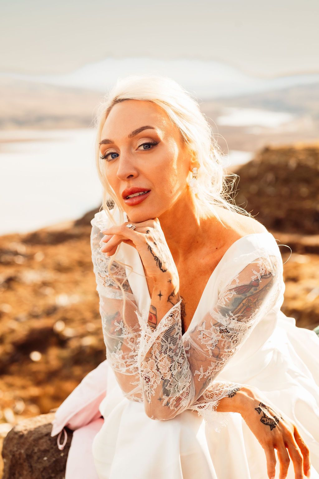 Blonde bride in a lace long-sleeve wedding dress poses outdoors with a scenic landscape behind her.