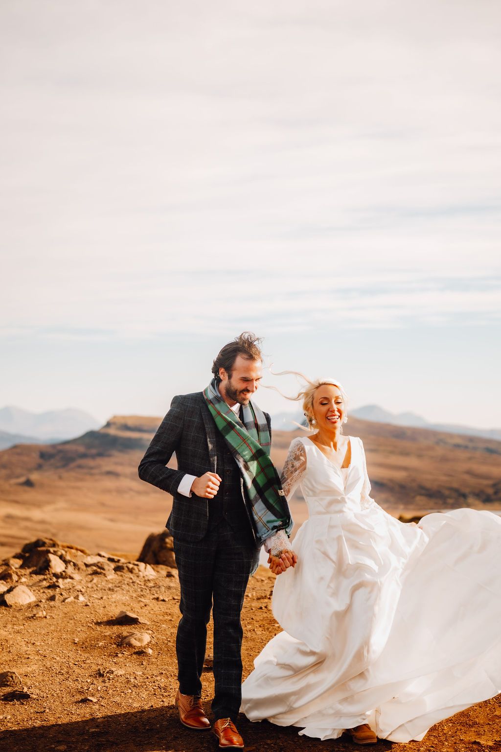 Laughing bride and groom walking hand in hand on windswept highland landscape under cloudy sky.