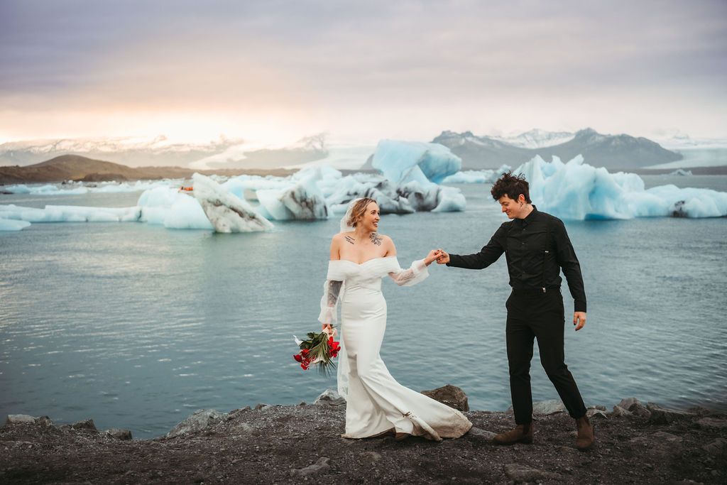 Bride and groom holding hands at Iceland glacier lagoon with blue icebergs at sunset.