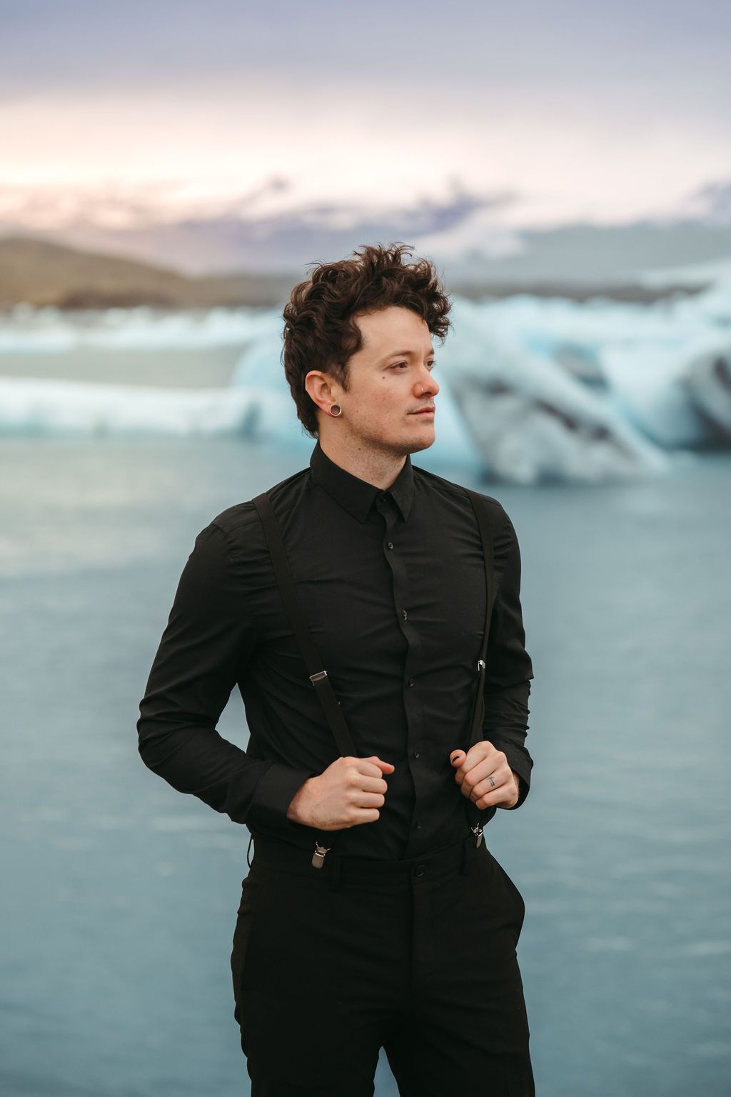 Man in black shirt and suspenders standing before glacial lagoon at dusk in Iceland.