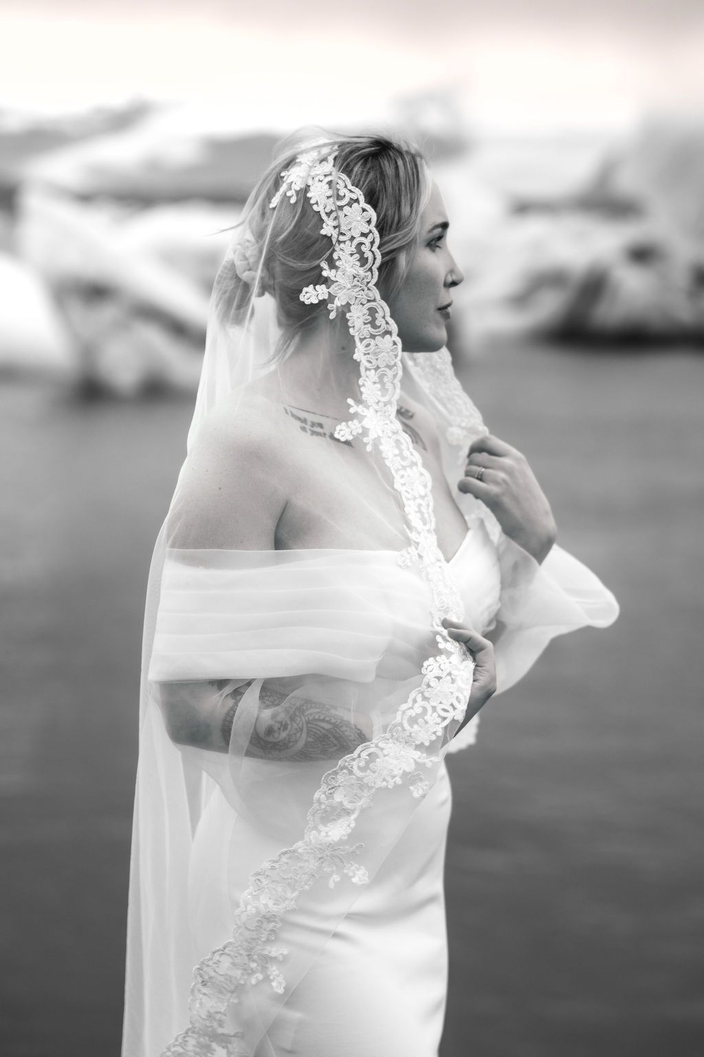 Black and white portrait of a bride in a white gown and lace veil standing near a body of water.