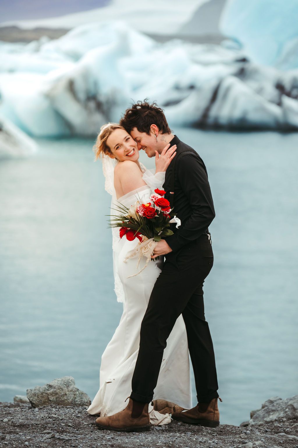 Bride and groom sharing a tender moment with red bouquet before a stunning glacial lagoon backdrop.