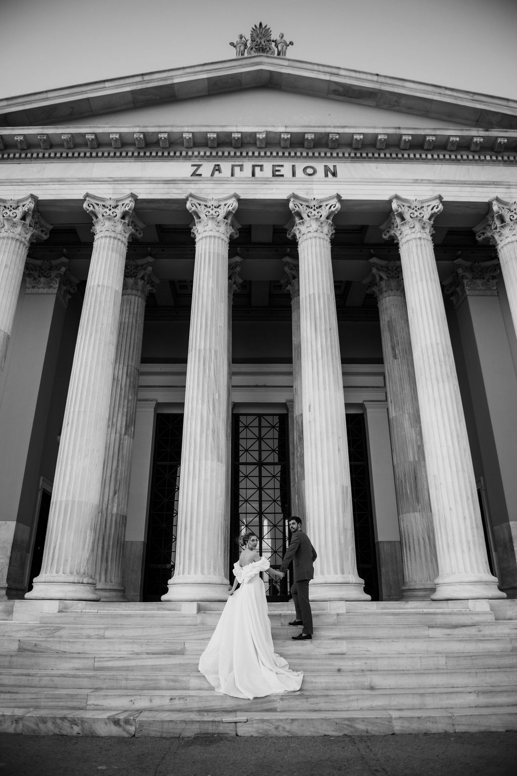 Bride and groom pose on steps of Zappeion neoclassical building in Athens, Greece, in elegant black and white wedding photo.