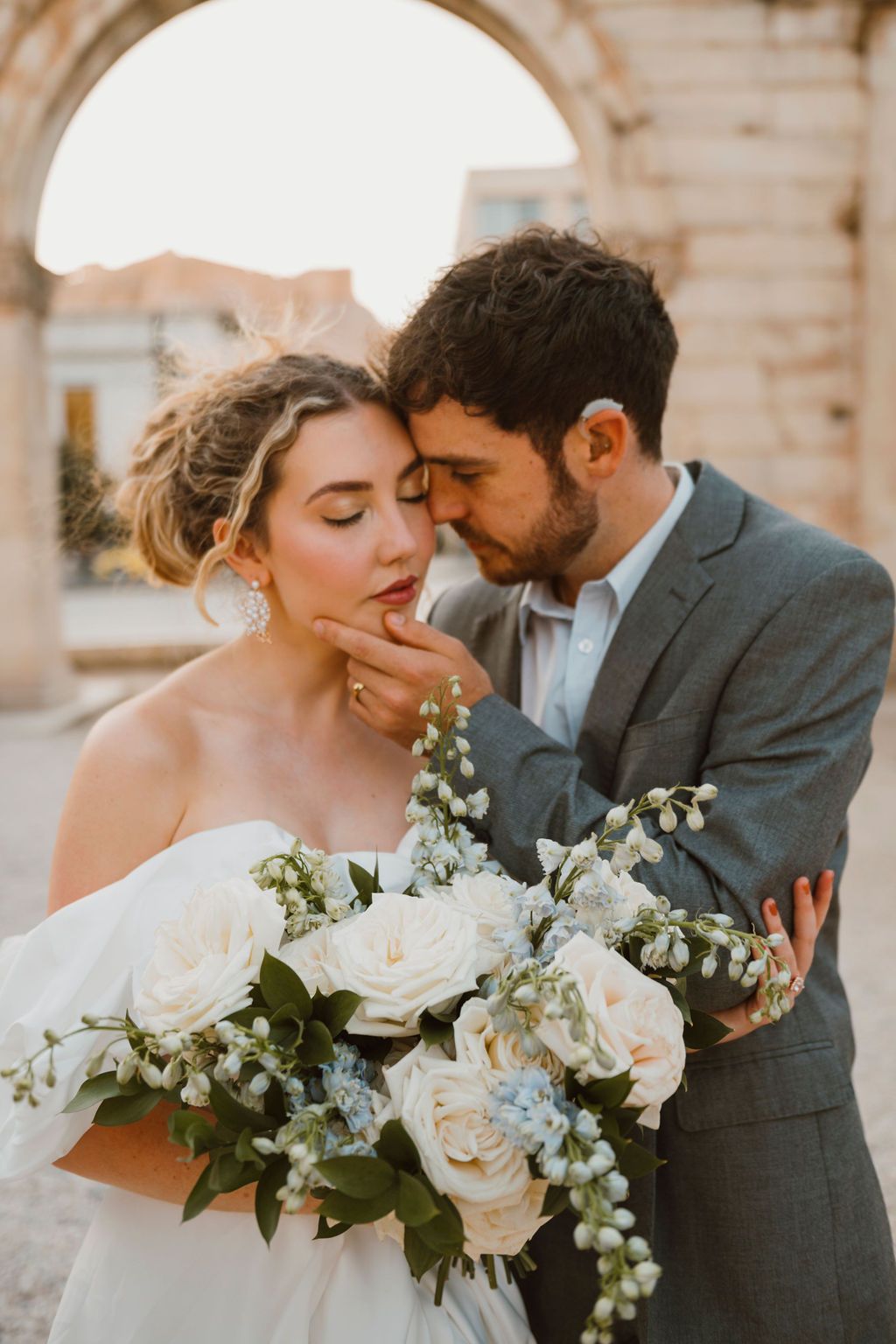 Bride and groom share a romantic moment, bride holding a white and blue floral bouquet near an arch.