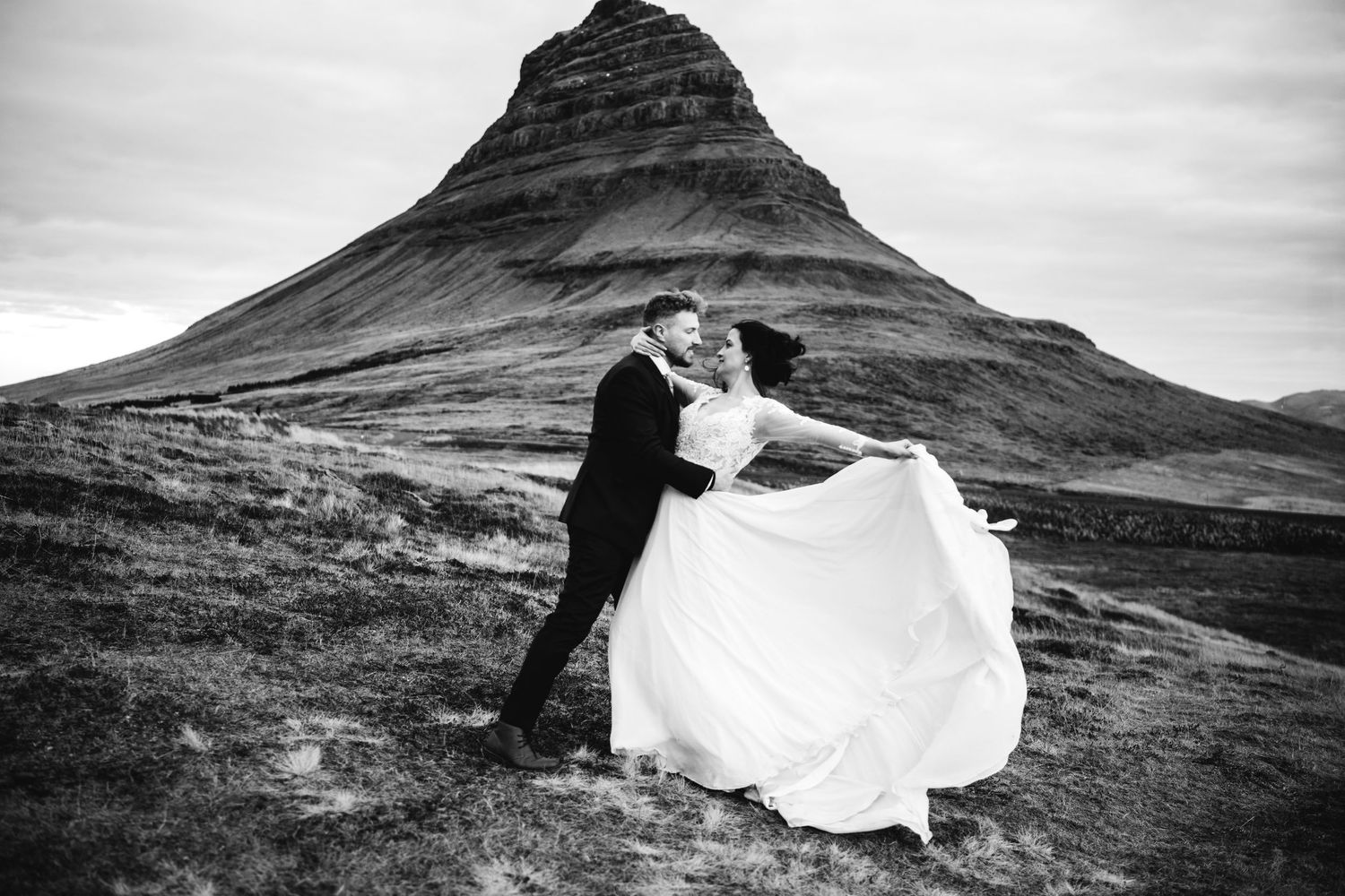 Black and white wedding photo of couple kissing before Kirkjufell mountain in Iceland, bride's dress flowing in wind.