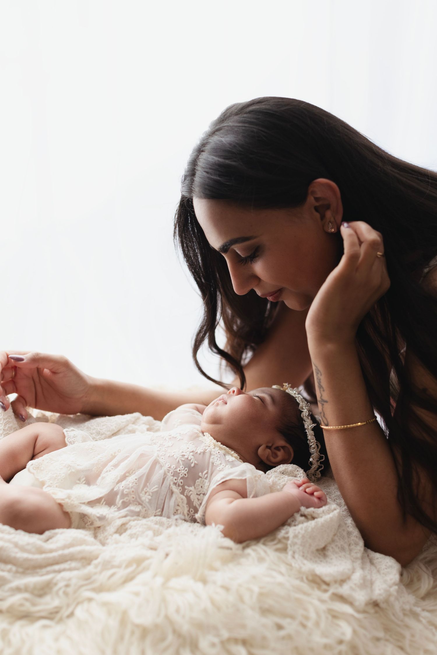 A mother with long dark hair leans over her newborn baby in a white lace outfit on a fluffy cream blanket.
