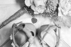 Black and white wedding detail photo featuring bridal shoes, a heart charm bracelet, and a floral bouquet.