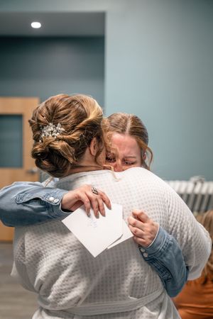 Two women share an emotional hug, one in a wedding updo with hair accessories, holding a note at their wedding day.