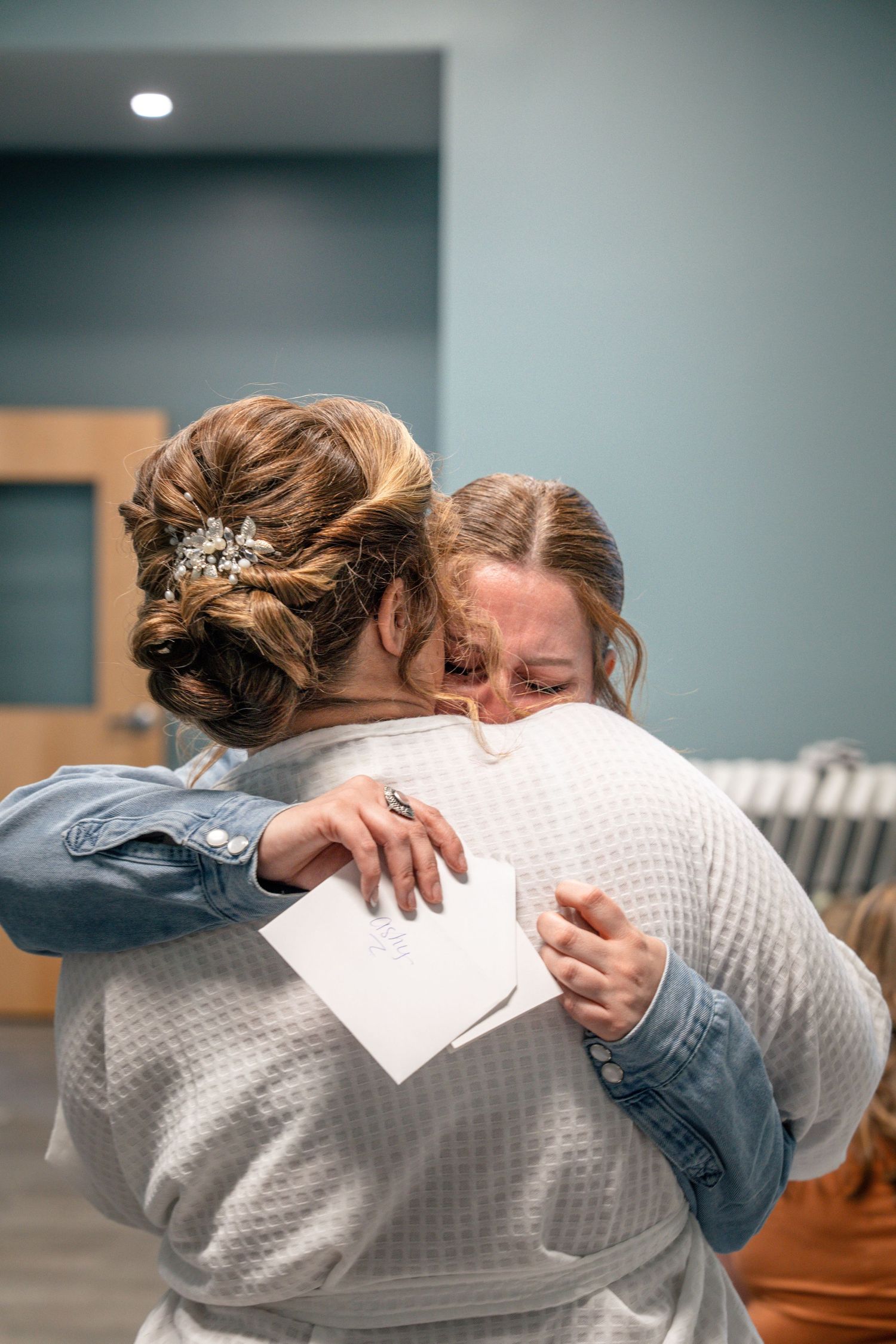 Two women share an emotional hug, one in a wedding updo with hair accessories, holding a note at their wedding day.