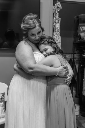Black and white photo of a bride in a white gown sharing a tender hug with a young girl in a formal dress.