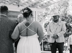 Black and white outdoor wedding ceremony with bride in backless gown, groom in light suit, and officiant under pergola.