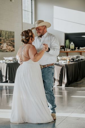 Bride and groom share first dance at rustic wedding reception, groom wearing cowboy hat, jeans, and white shirt.