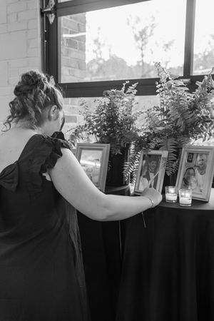 Woman in black dress viewing memorial table with framed photos, candles, and greenery in black and white photo.