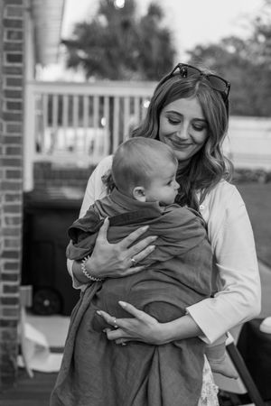 Black and white photo of a smiling woman holding a baby in a wrap carrier outdoors near a brick building.