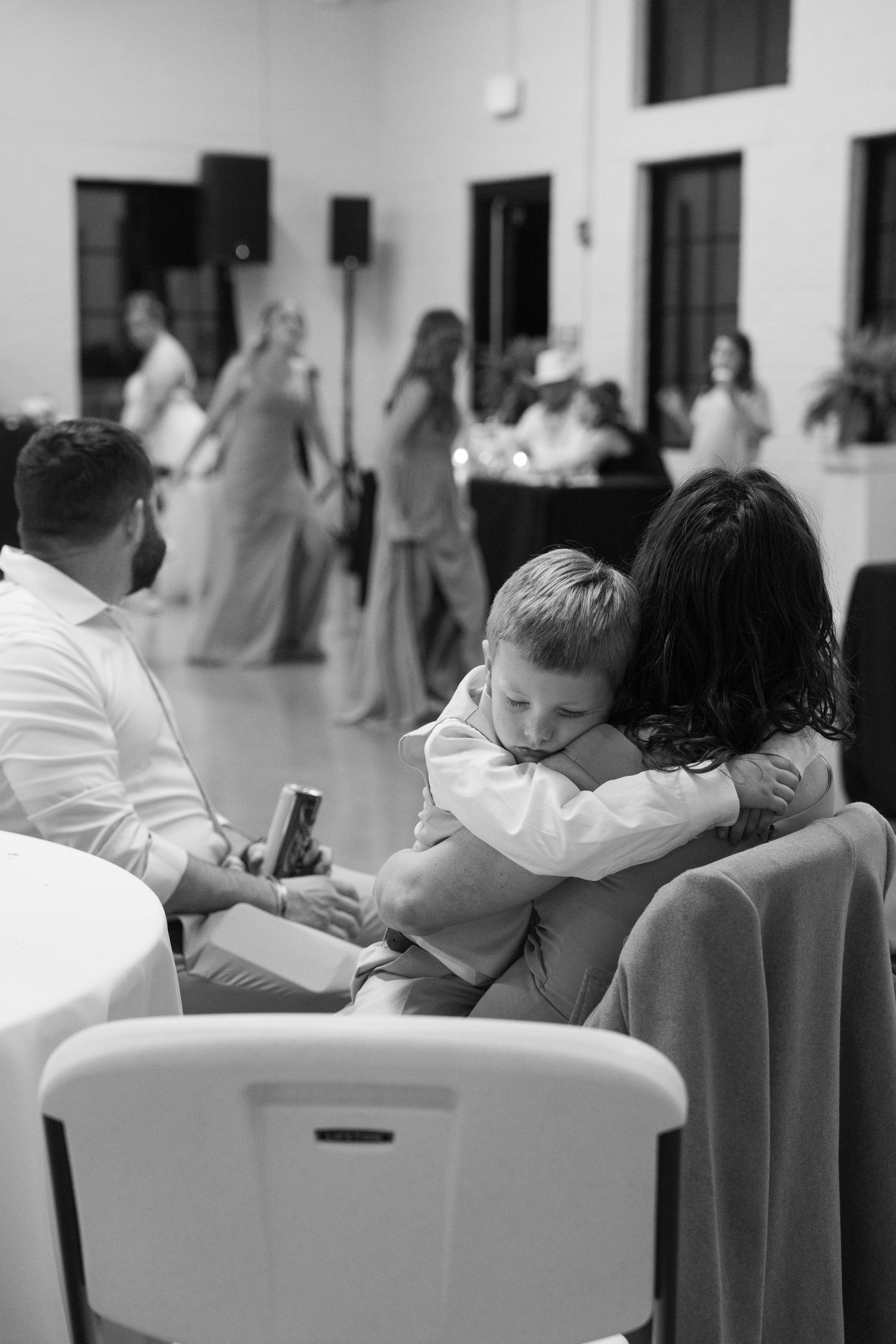 A young boy in a suit hugs a woman at a wedding reception while guests dance in the background.