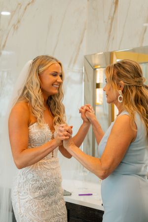 Bride in lace gown and veil holds hands with woman in blue dress, sharing a joyful moment in a marble bathroom.