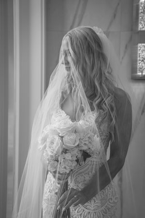 Black and white photo of a bride in lace gown and veil holding a white rose bouquet, gazing down elegantly.