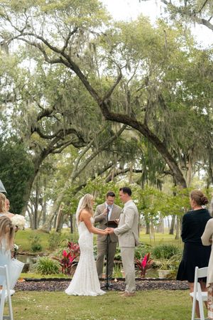 Outdoor wedding ceremony under Spanish moss-draped oak trees, bride in white gown and groom in tan suit exchanging vows.