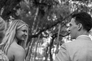 Black and white photo of a smiling bride and groom exchanging vows outdoors among trees.