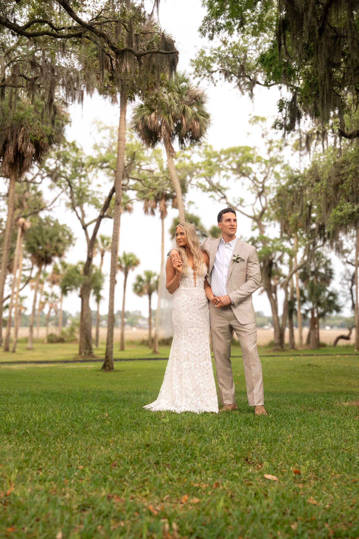 Bride and groom stand together on lush green lawn surrounded by Spanish moss-draped oak and palm trees.
