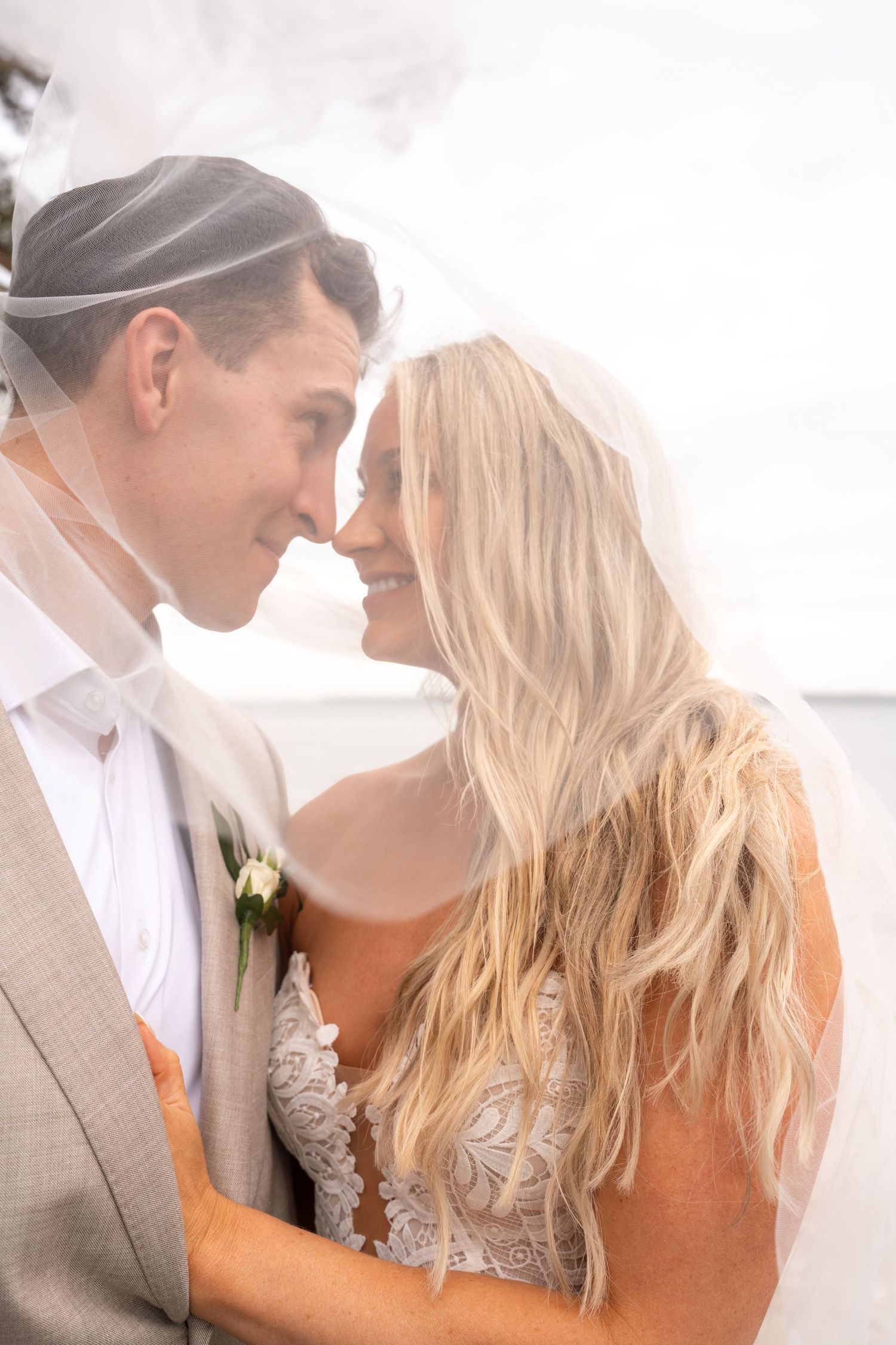 Bride and groom share a romantic moment under a flowing veil, touching foreheads and smiling on their wedding day.