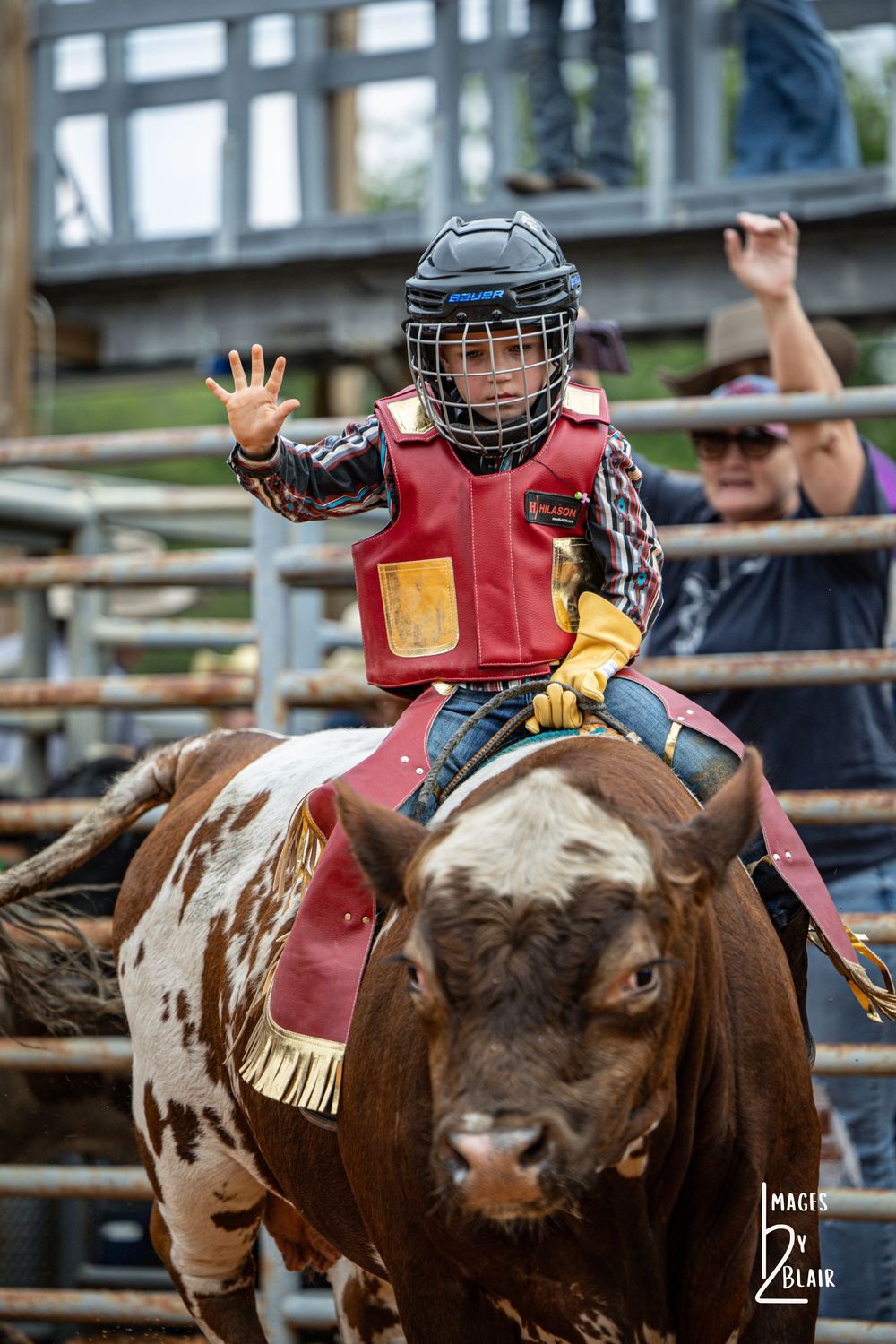 Young child in protective gear riding a bull at a rodeo event, waving hand while wearing a helmet and red vest.