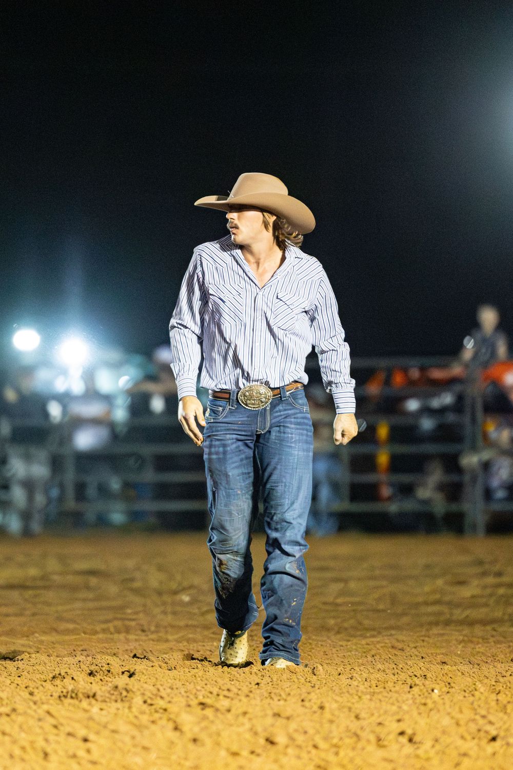 Cowboy in striped shirt, jeans, and hat walks across a rodeo arena dirt floor at night under bright lights.