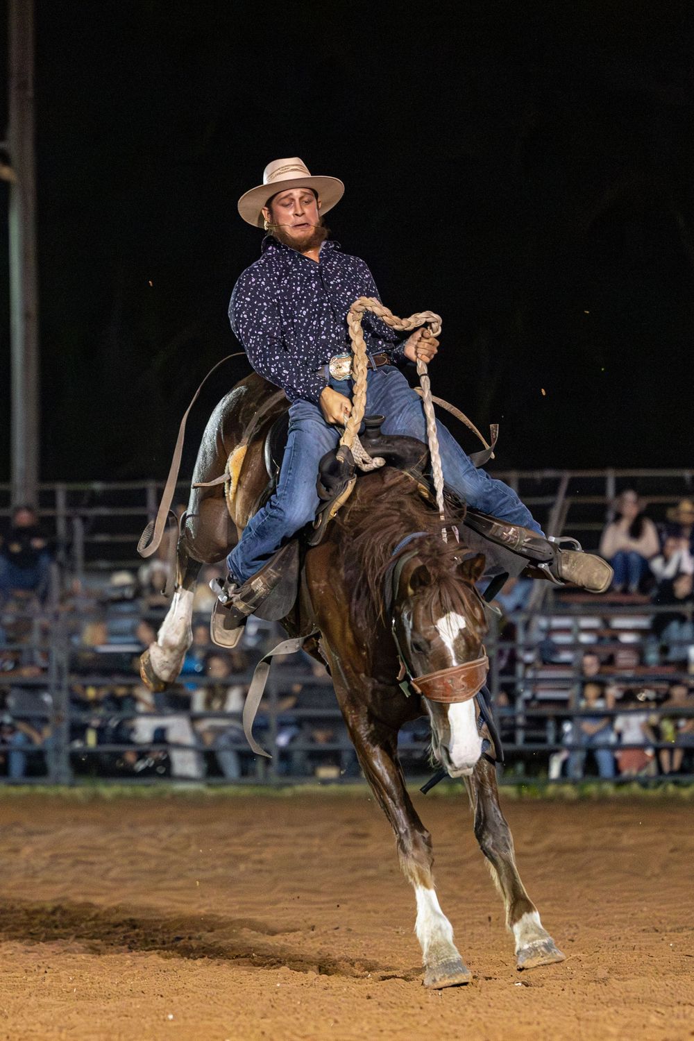 Cowboy in hat rides bucking bronco at night rodeo, gripping rope tightly in blue shirt and jeans.