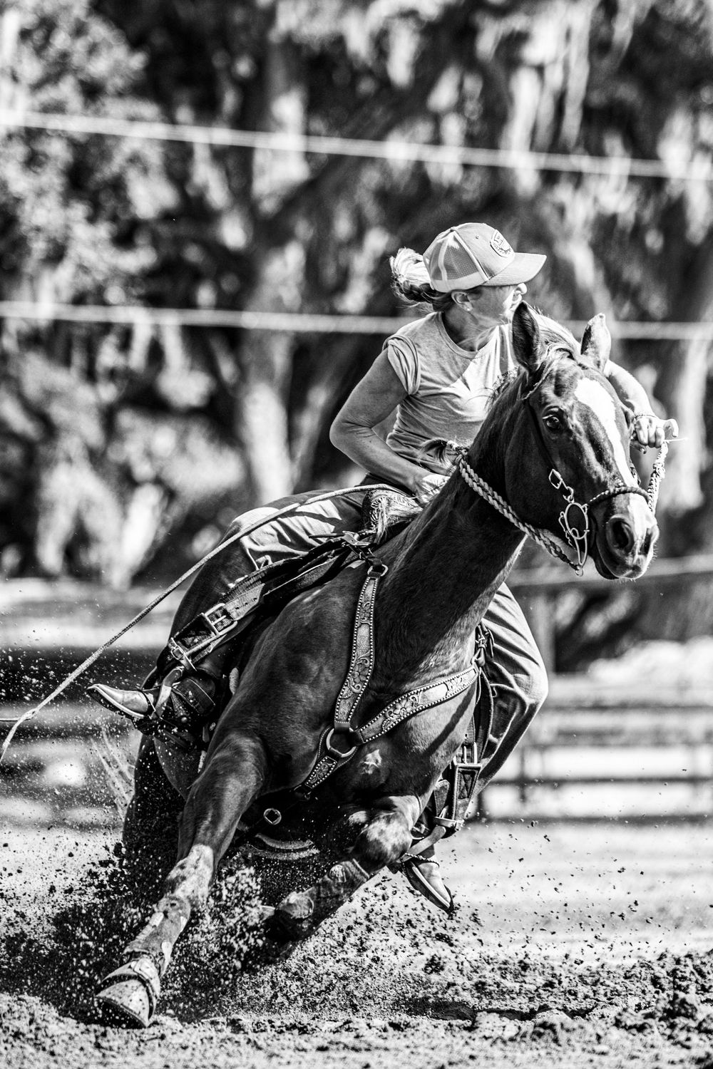 Black and white photo of a female rider racing on horseback through dirt at a rodeo barrel racing competition.