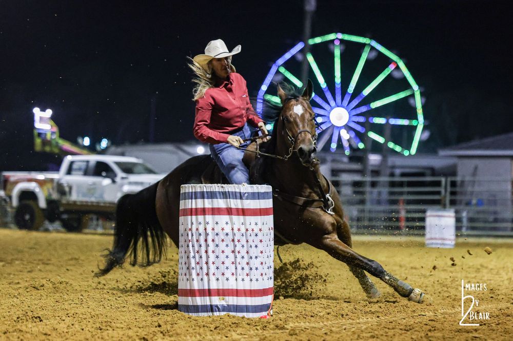 Woman in pink shirt barrel racing on dark horse at night rodeo with Ferris wheel lights in background.